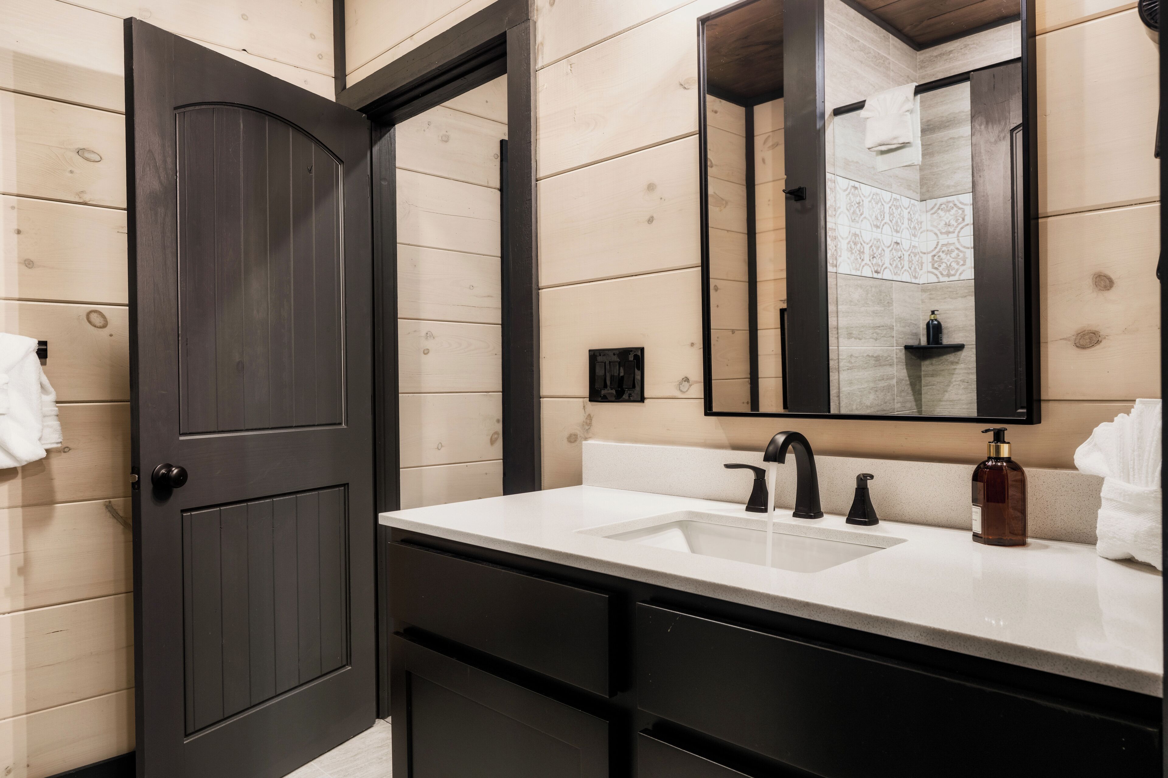 A modern bathroom with a dark wooden door, a white sink, black faucets, and a large mirror.
