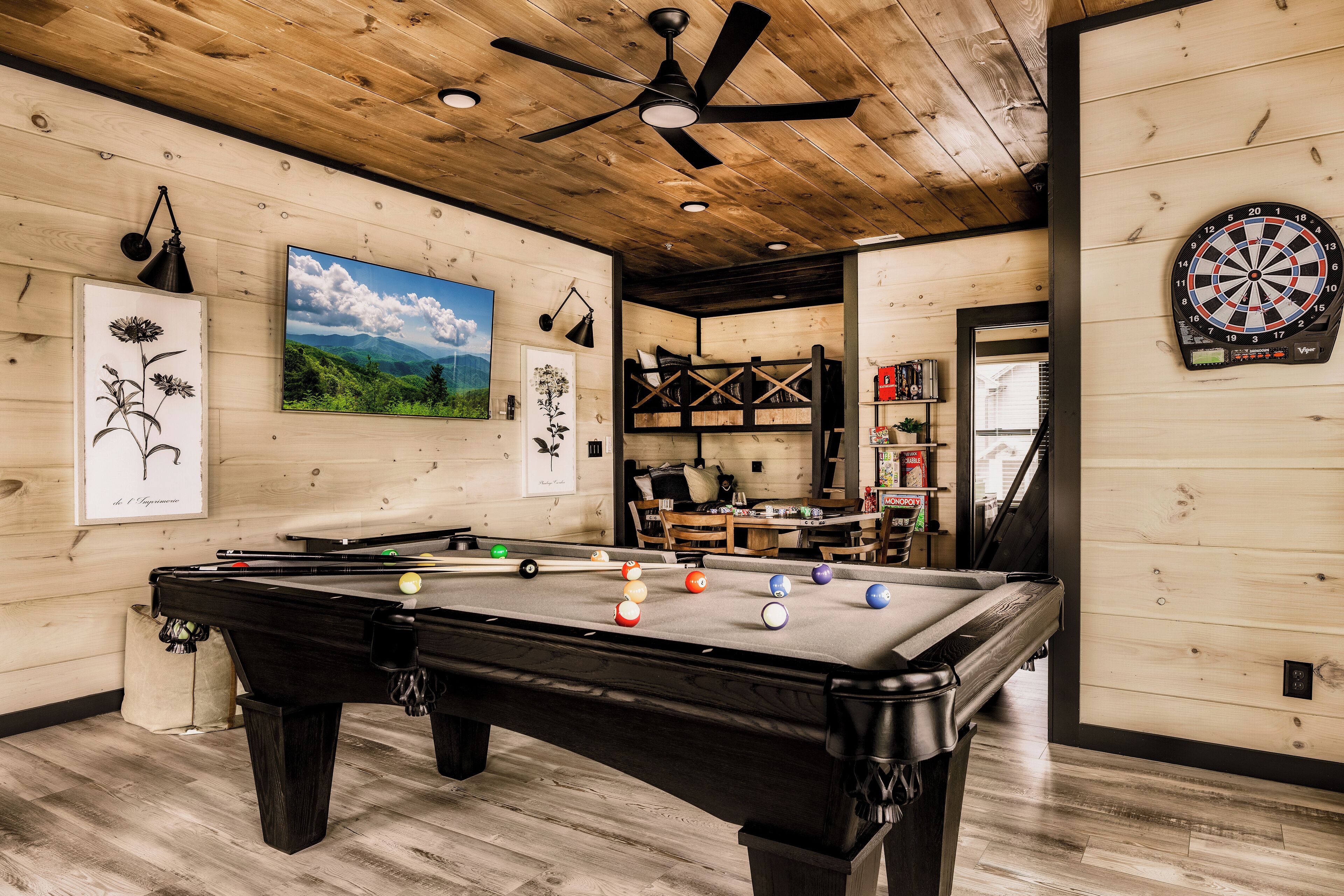 A pool table with balls, a dartboard, and a ceiling fan in a room with wooden walls and a wooden ceiling.