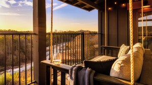 A wooden deck with a swing seat, a glass on the railing, and a view of a road and trees.