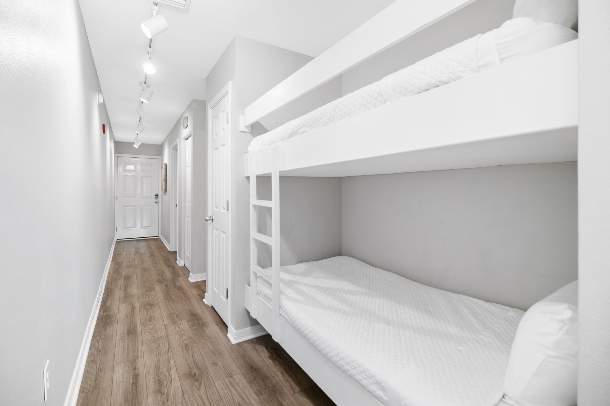 A bunk bed room with white bedding, wooden flooring, and a white door at the end of the hallway.