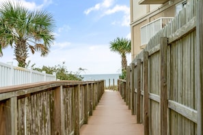 A wooden boardwalk leading to a beach, flanked by wooden fences and palm trees.