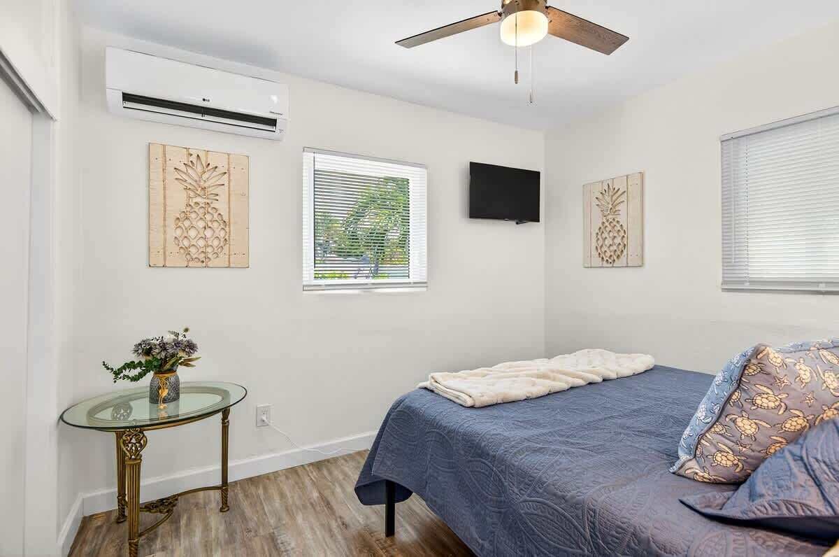 Een slaapkamer met een bed, een glazen tafel met een vaas, een plafondventilator en een aan de muur bevestigde televisie.