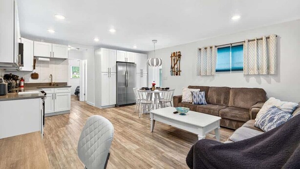 Eine moderne Küche mit weißen Schränken, ein Essbereich mit weißem Tisch und Stühlen, ein braunes Sofa und ein großes Fenster mit Vorhängen.