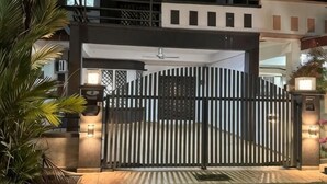 Uma entrada murada com um portão de metal, uma palmeira e um edifício com varanda ao fundo.