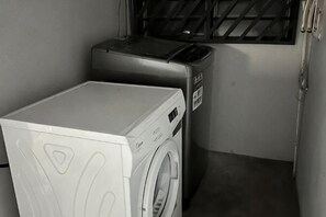 A compact laundry area with a washing machine and dryer, a window with bars, and a door with a handle. - JL Homestay Bahau (Bahau)