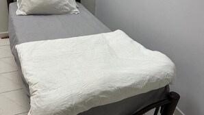 Ein Einzelbett mit grauem Matratze und weißer Bettwäsche, ein hölzernes Kopfteil und eine schlichte Wand.