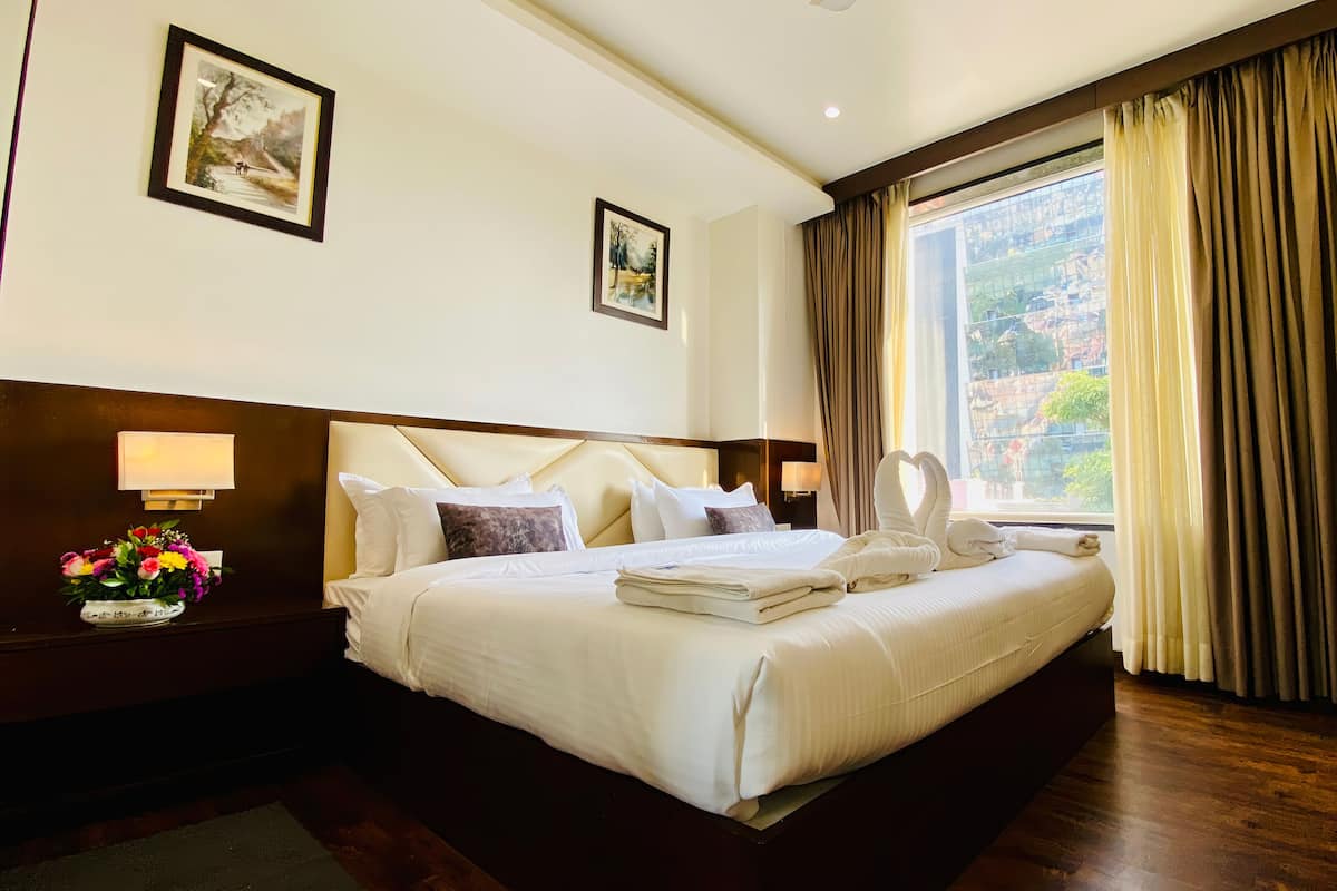 A neatly made bed with white linens and pillows, a bedside table with a lamp and a vase of flowers, and a large window with curtains.