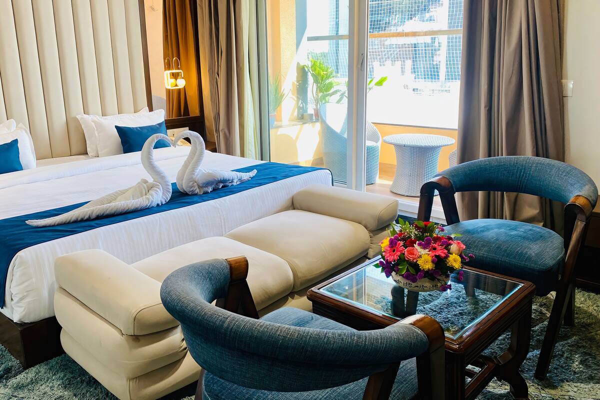 A hotel room with a bed, two armchairs, a glass coffee table with a floral centerpiece, and a view of the outdoors through large windows.