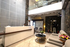 Zona de recepción de hotel con mostrador, área de descanso con sofá y mesa de centro, y una pared con exhibición de objetos.