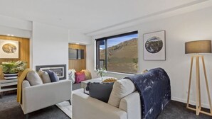 A modern living room with a fireplace, a television, and a large window with a mountain view. - Residence Du Parc - Awa Apt 16a (Arthurs Point)