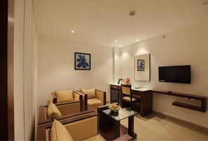 A modern hotel room with a sofa set, a coffee table, a flat-screen TV mounted on the wall, and a small desk with a chair.
