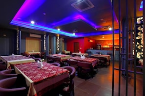 A restaurant with tables covered in red tablecloths, a bar area in the background, and purple lighting on the ceiling.
