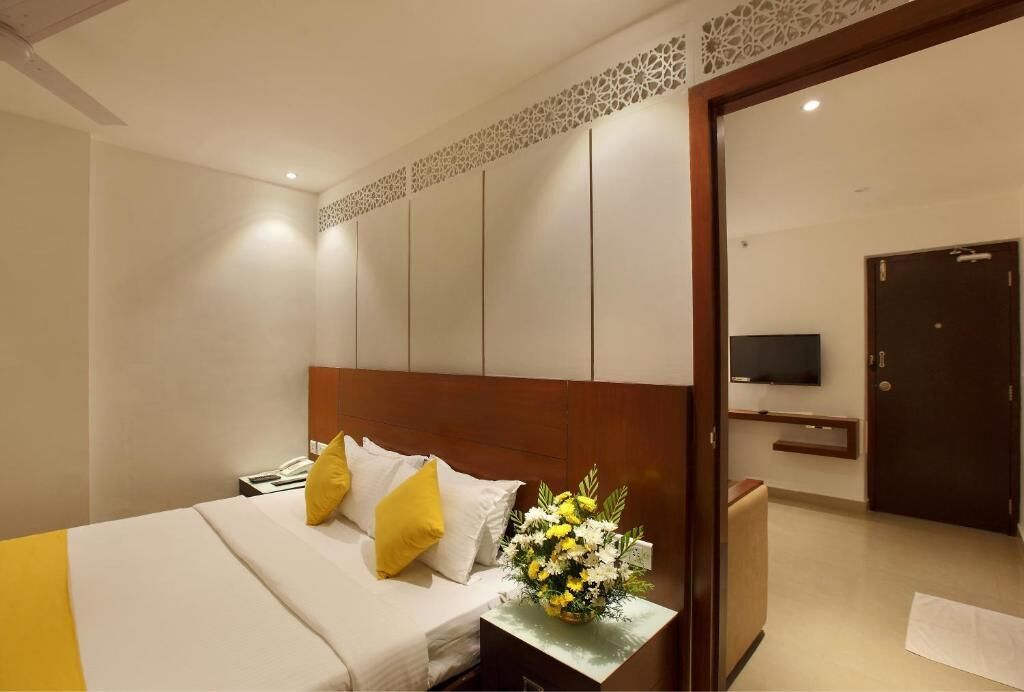 Ein Hotelzimmer mit einem Bett, einem Nachttisch mit Blumen in einer Vase, einem an der Wand montierten Fernseher und einer Tür rechts.