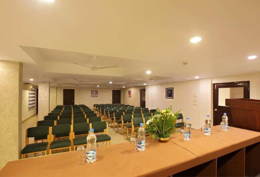Un salón de conferencias con filas de sillas verdes, una larga mesa con botellas de agua y un ventilador de techo.