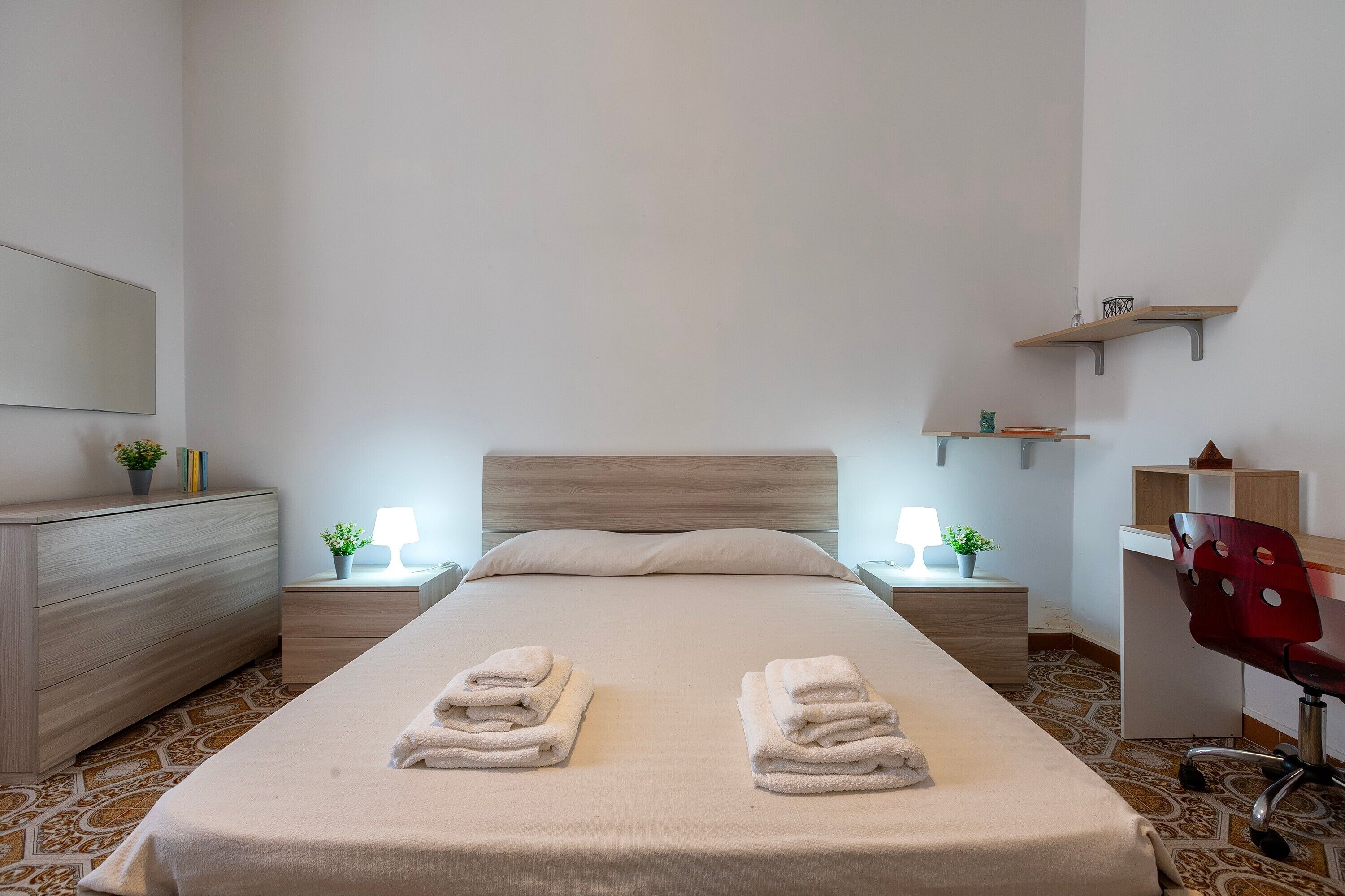 A neatly made bed with two sets of folded towels, a wooden dresser, bedside tables with lamps, and a desk with a chair.