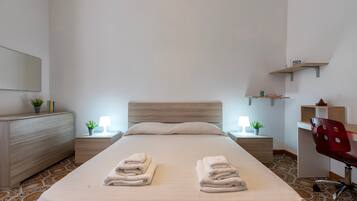 A neatly made bed with two sets of folded towels, a wooden dresser, bedside tables with lamps, and a desk with a chair.