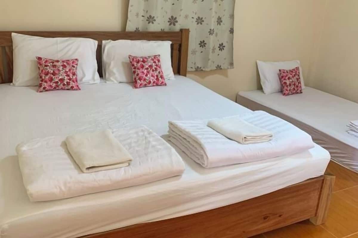 Un grand lit à deux places avec du linge de lit blanc et des coussins à motifs floraux roses, un lit simple plus petit avec du linge de lit similaire, et des serviettes soigneusement pliées posées sur le lit.