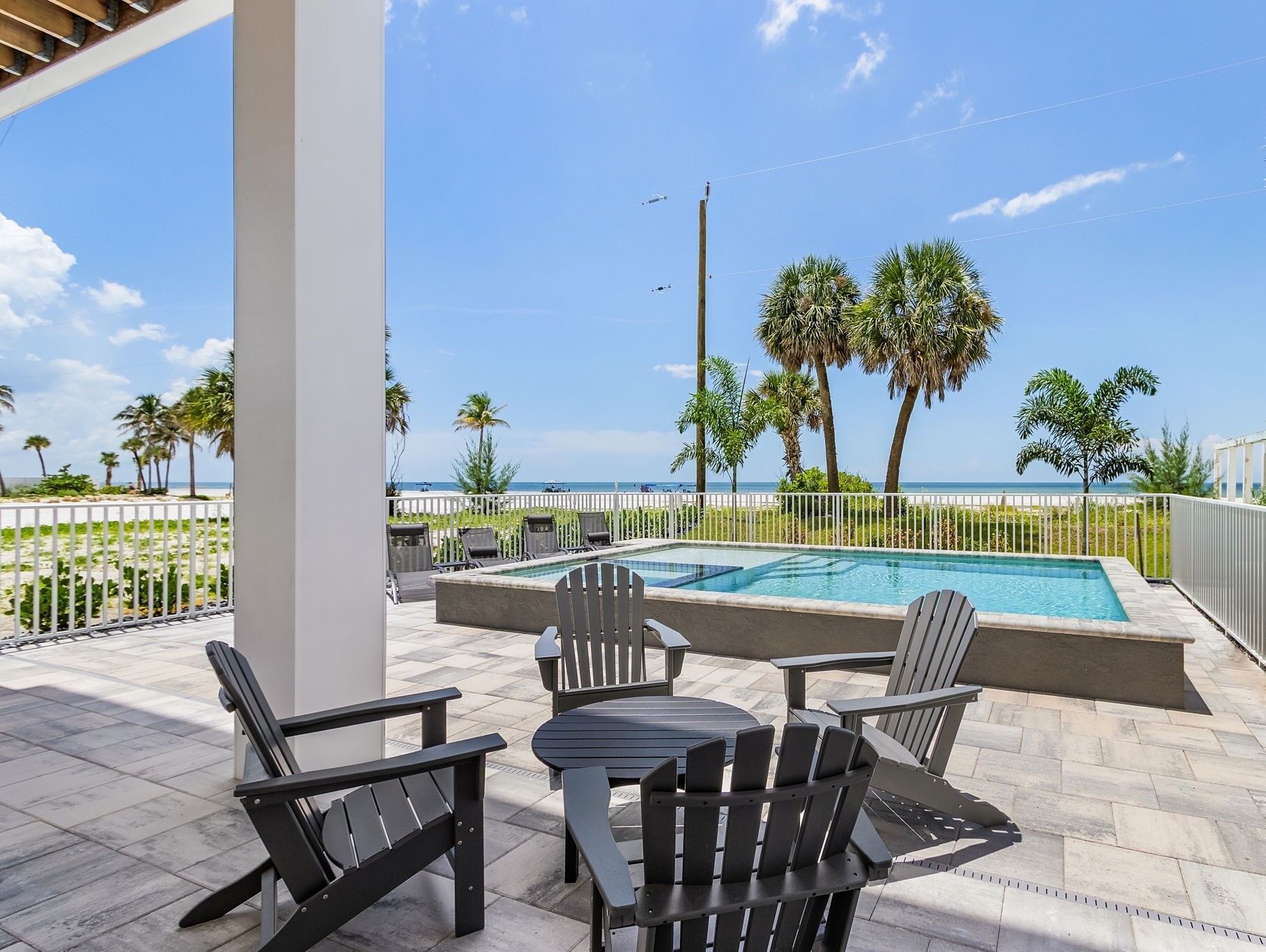 A pool area with a view of the beach, outdoor seating, and palm trees.