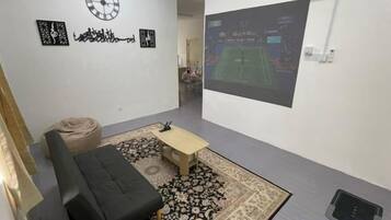 A room with a black sofa, a small table, a patterned rug, a wall clock, and a projection screen displaying a tennis match.