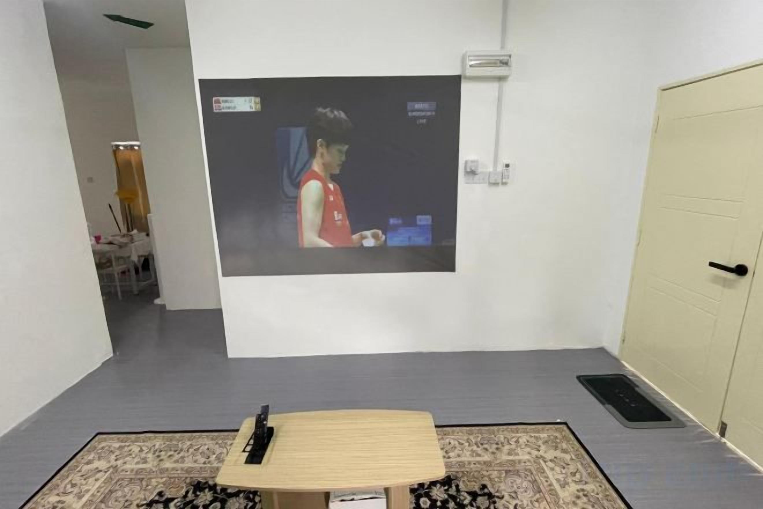 Een kamer met een projectiescherm waarop een persoon staat afgebeeld, een tafel met een afstandsbediening en een deur.