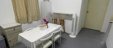 Una habitación pequeña y limpia con cama, mesita de noche y escritorio con dos sillas. Tiene una ventana con cortinas, una puerta y un radiador.