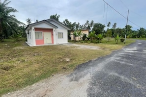 Une petite maison avec une porte rouge, entourée de pelouse et bordée d’une route goudronnée.