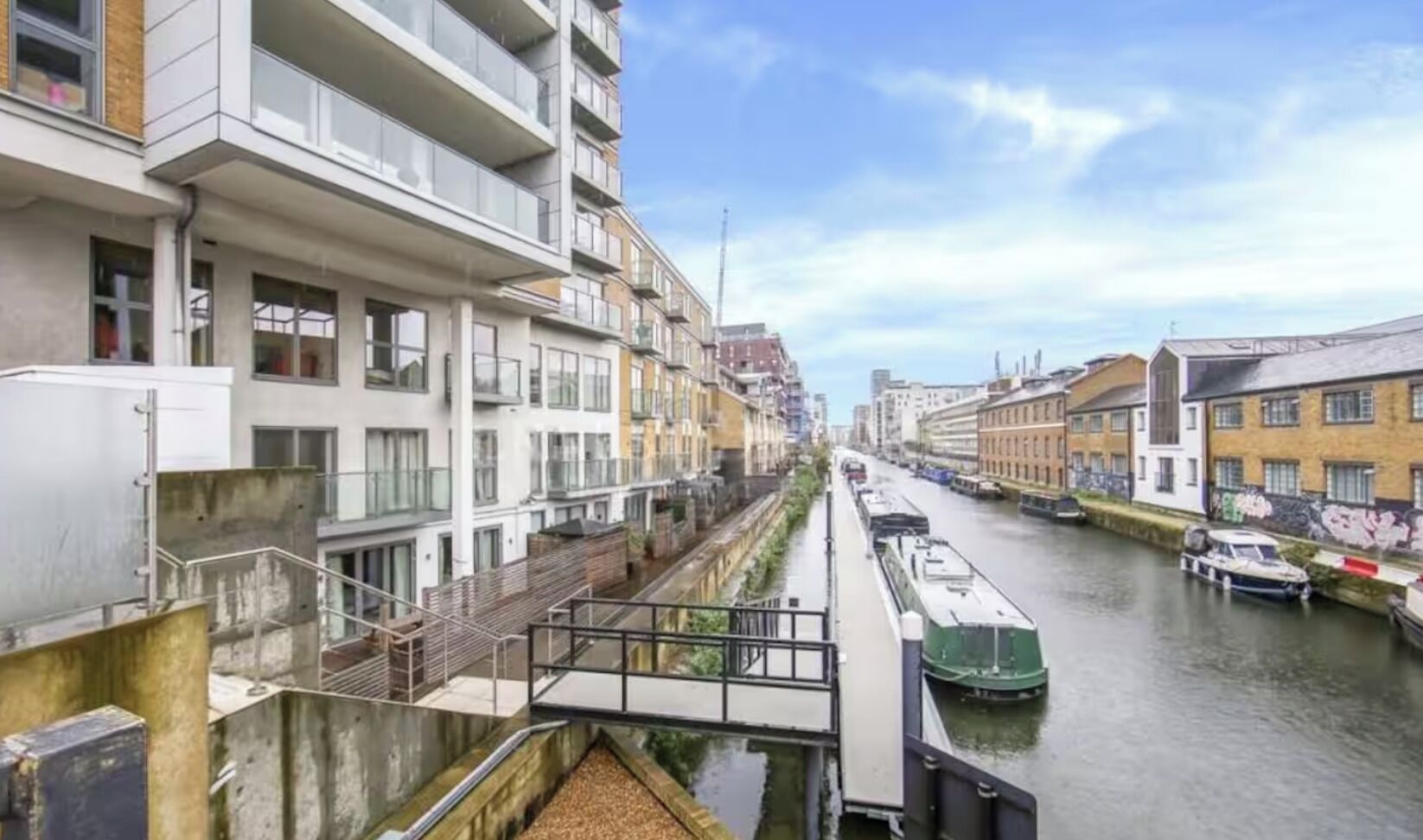 Vista de un moderno entorno urbano a orillas de un canal, con edificios de varios pisos, un puente peatonal y un tren verde.
