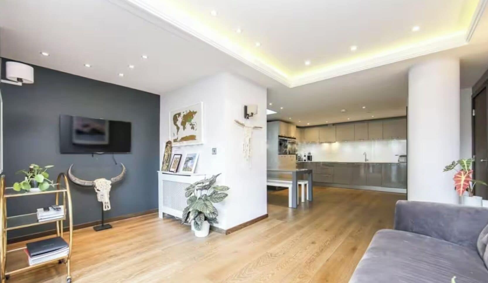 Una sala de estar moderna con un sofá gris, piso de madera, un televisor montado en la pared y una cocina al fondo.