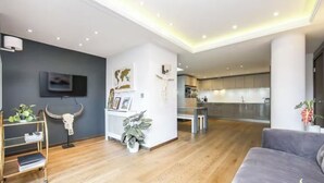 A modern living room with a grey sofa, wooden flooring, a TV mounted on the wall, and a kitchen in the background. - Spacious, Canal-Side, 3bed flat (London)