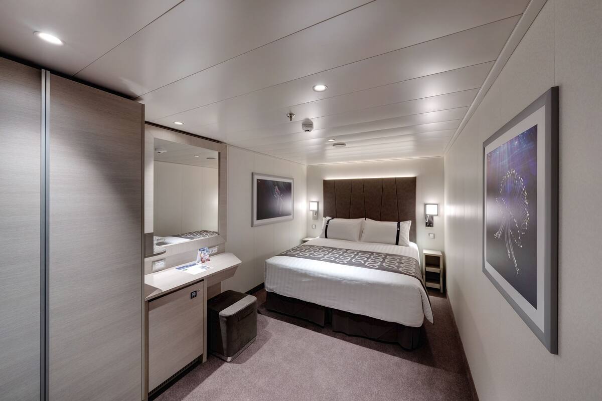 Ein modernes Hotelzimmer mit einem großen Bett, einem Schreibtisch, einem Sessel, einem kleinen Tisch, einem Spiegel und einem an der Wand befestigten Fernseher.