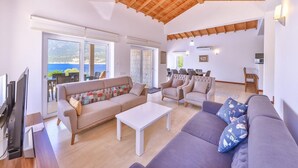 Ein geräumiger Wohnbereich mit hohen Holzbalken, einem beigen Sofa mit bunten Kissen, einem weißen Couchtisch und Meerblick durch große Fenster.
