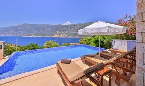 Piscina infinita con vistas al mar y a las montañas, sillones de madera y una sombrilla blanca.
