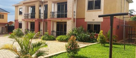 A two-story residential building with a paved courtyard, a small garden, and a metal fence.