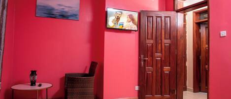 Une pièce avec un mur rouge, un téléviseur fixé au-dessus d’un fauteuil en osier, une petite table ronde avec un vase, et une porte en bois.