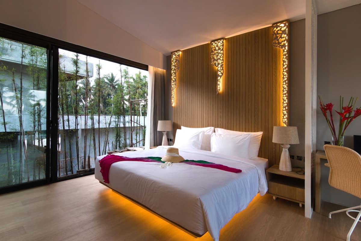 A modern hotel room with a large bed, a nightstand with lamps, a chair, and a view of palm trees through large windows.