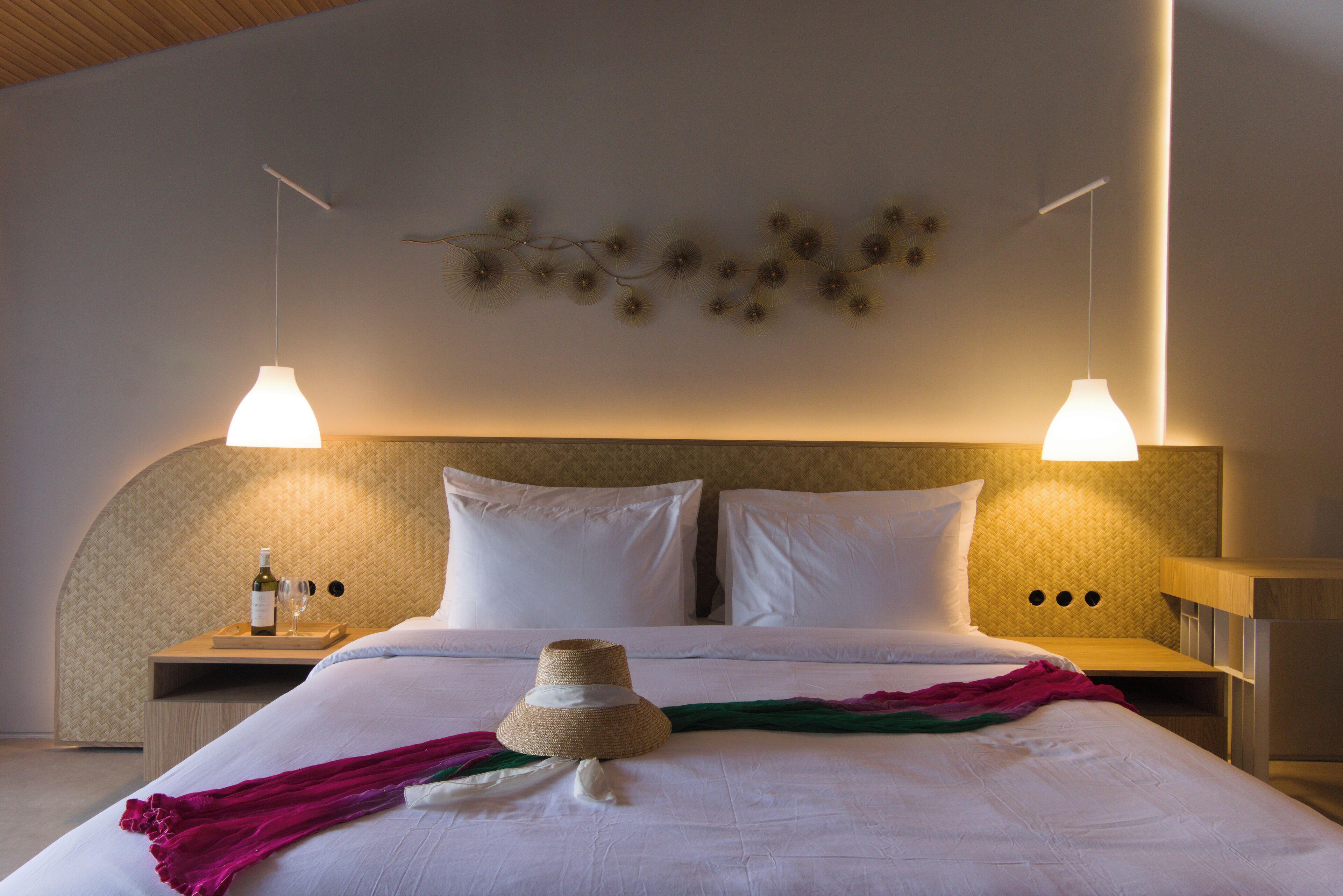 Een net opgemaakt bed met witte lakens, een geweven hoed en een fles wijn op het nachtkastje.