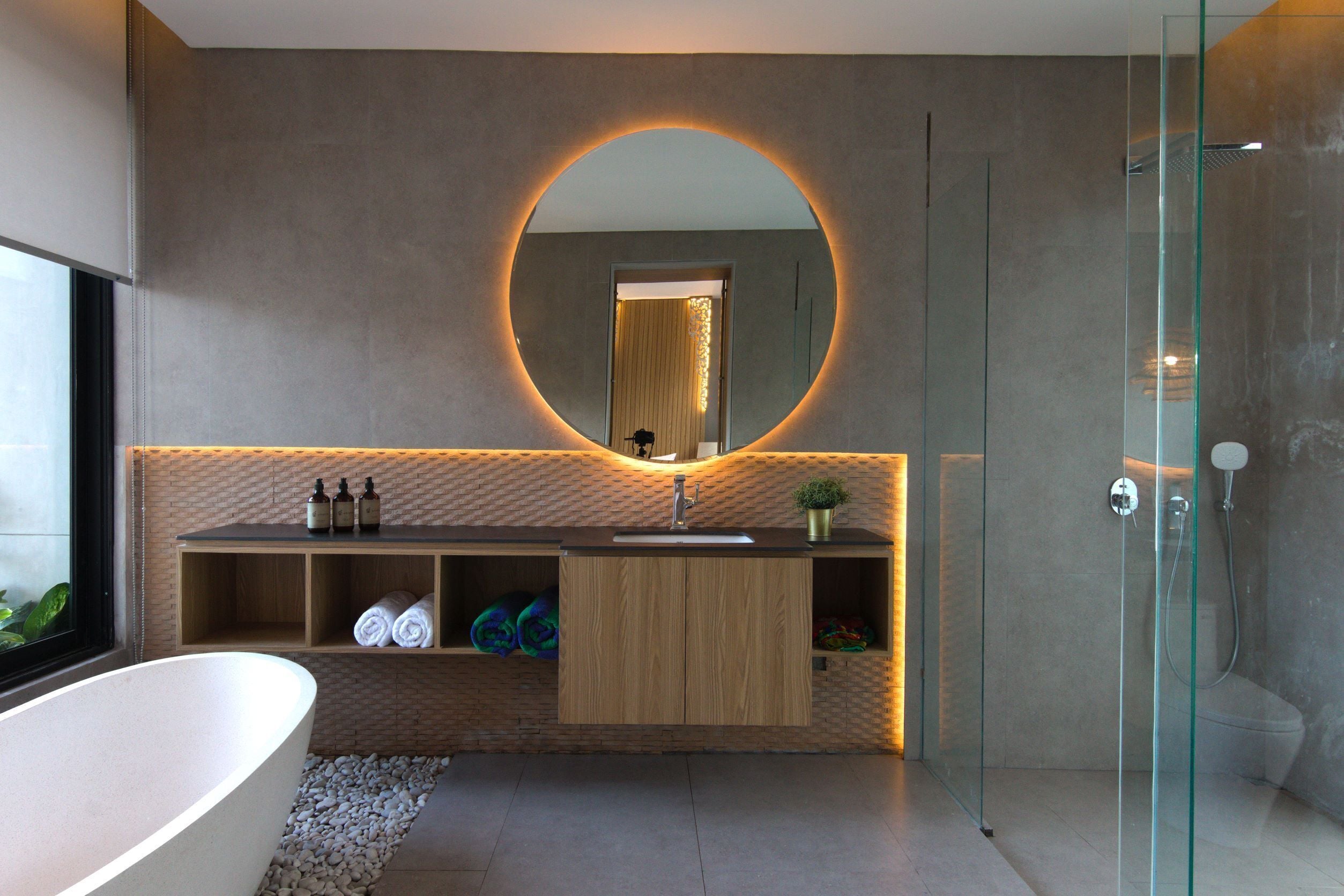 Un baño moderno con una bañera independiente, un espejo redondo con luz circular, una cabina de ducha de vidrio y un mueble de tocador de madera con estantes integrados.