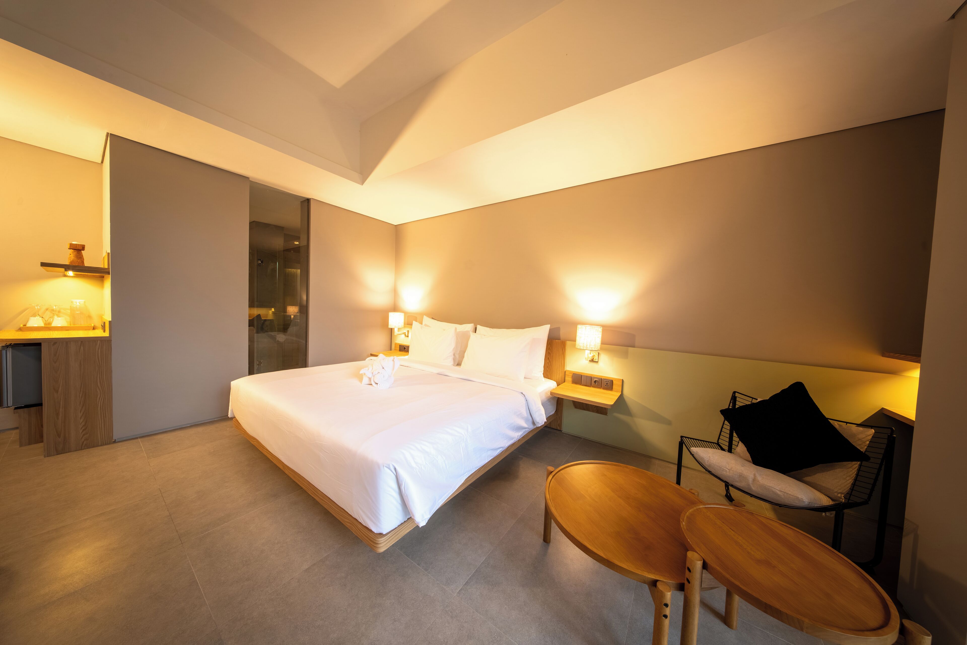 Quarto de hotel moderno com uma cama grande, mesas de cabeceira com luminárias, uma pequena mesa de madeira e uma poltrona com almofadas.