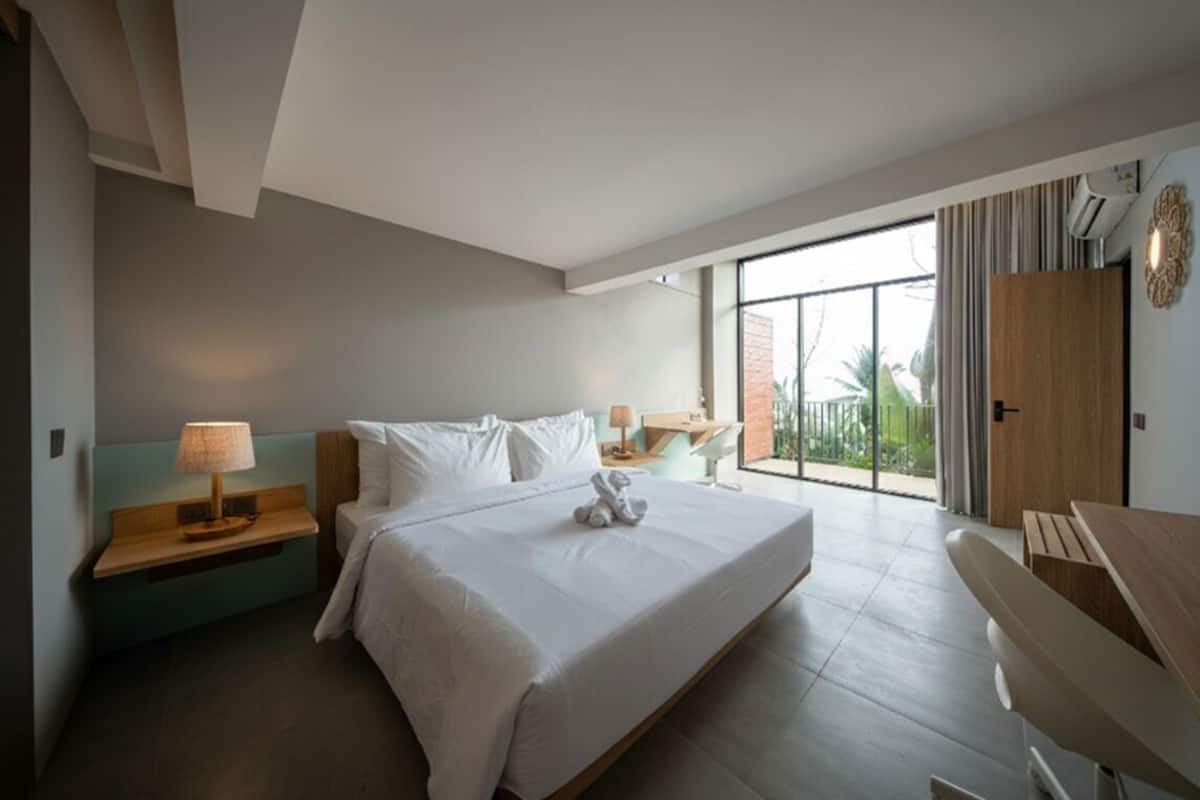 A modern hotel room with a large bed, a bedside table with a lamp, a wooden bench, and a view of the outdoors through a large window.