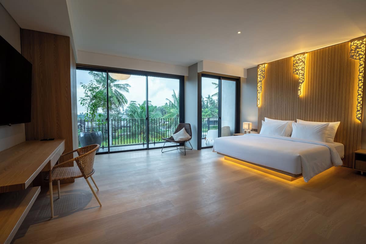 A modern bedroom with a large bed, a desk, and a chair. There is a balcony with a view of palm trees.
