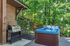 Une cabane en bois dotée d’un barbecue et d’un bain à remous sur une terrasse en bois, le tout entouré d’arbres.
