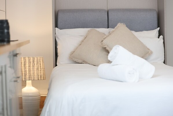 Una cama bien hecha con sábanas blancas y dos almohadas color beige, una lámpara blanca con pantalla a rayas, y una toalla blanca enrollada sobre la cama.
