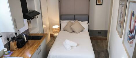 A compact hotel room with a bed, a bedside table, a small kitchenette, and a bathroom.