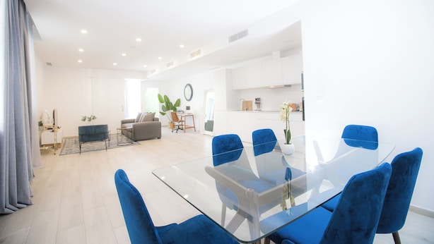 Uma sala de estar moderna com uma mesa de jantar de vidro e cadeiras azuis, um sofá e uma mesa de centro com uma planta.