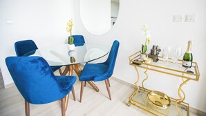 Uma área de refeições moderna com mesa de vidro, cadeiras de veludo azul, espelho com moldura dourada e uma mesinha lateral com moldura dourada, com uma garrafa e copos.
