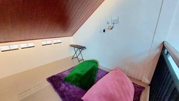 Una habitación con techo inclinado, una mesita pequeña, un tapete verde y rosa, y una pared blanca con un interruptor de luz.