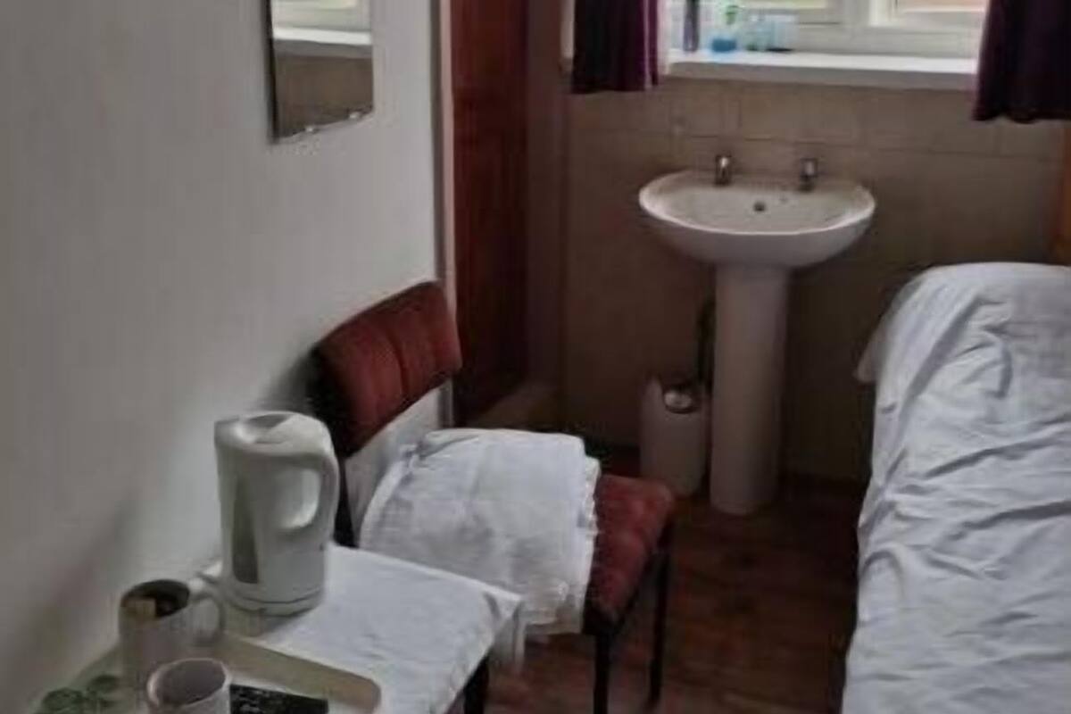 A small, simple room with a bed, a table with a kettle and cups, a chair, a sink, and a window with curtains.