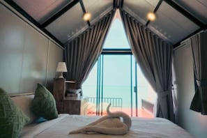 Un dormitorio con cama, almohadas, una mesita de noche, una lámpara y vistas al mar a través de un ventanal grande.