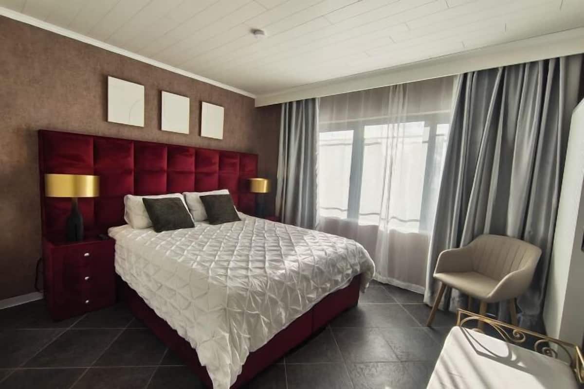 A hotel room with a large bed, a red headboard, two bedside lamps, a chair, and a large window with curtains.