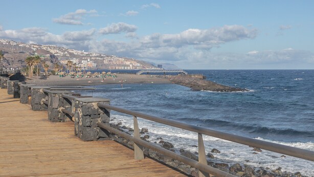Un muelle de madera con barandillas de piedra que da a una playa con gente y palmeras.